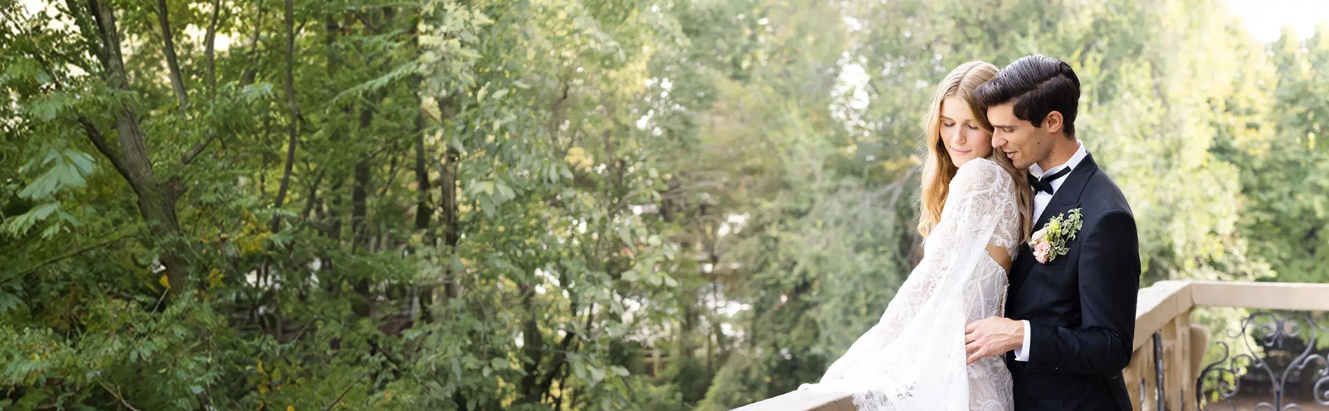 A bride and groom holding each other on a wooded terrace, in soft daylight.